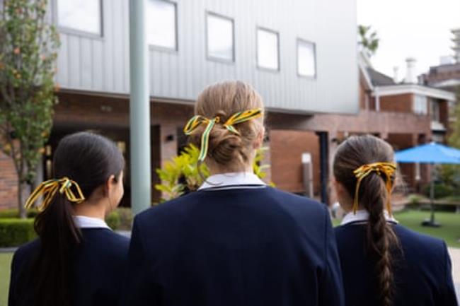 Three girls in dark blue school uniforms seen from behind with green and gold ribbons in their hair