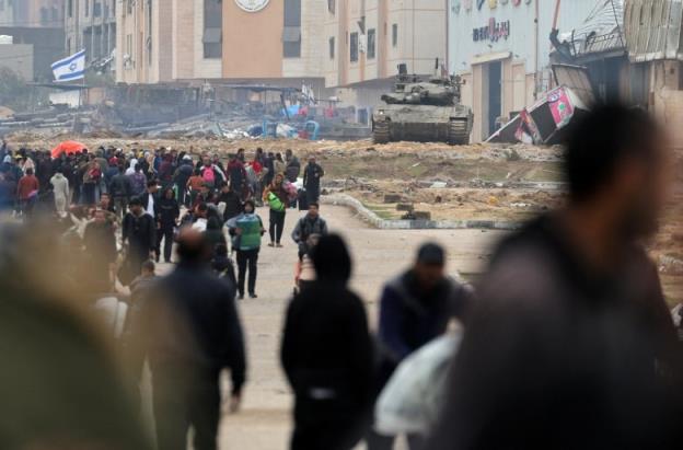 People walk down a road as a tank is seen in the background.