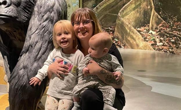 A woman embraces a baby and a toddler in front of a statue of a gorilla  in a museum.