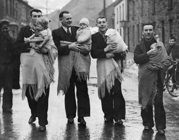 A black and white archival photos of men in suits walking with babies wrapped in shawls.