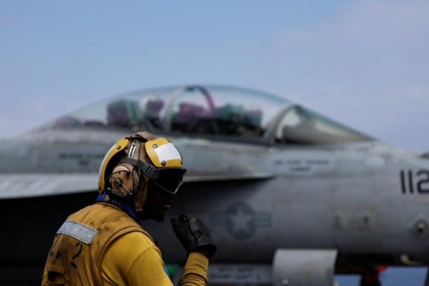 A person with their face covered and in a yellow suit and helmet holds a hand to their face. A fighter jet with the pilot in the closed cockpit is visible in the background.