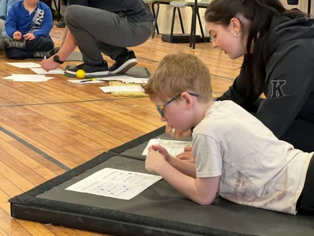 A boy with glasses sits on a mat and plays bingo.