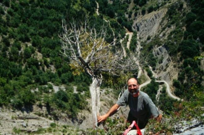 View from above of Doug Larson on a cliff holding a tree with a long drop behind him with a switchback road seen snaking round the mountain.